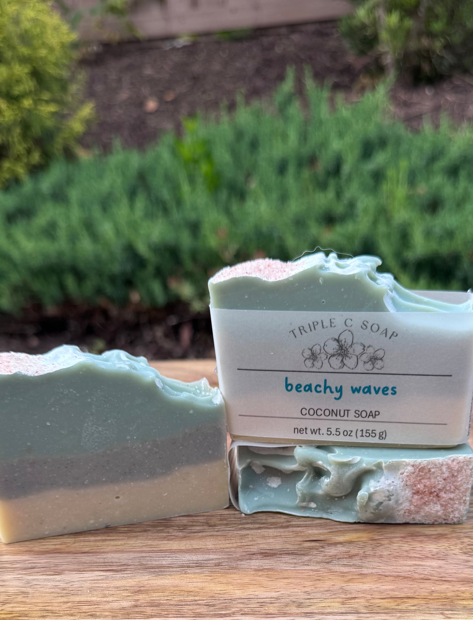 Coconut soap bar with 'Triple C Soap' packaging on a wooden surface with greenery in the background