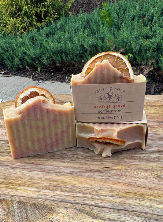 Three bars of soap with orange slices on a wooden surface, with label 'Orange Grove' in front of natural background.