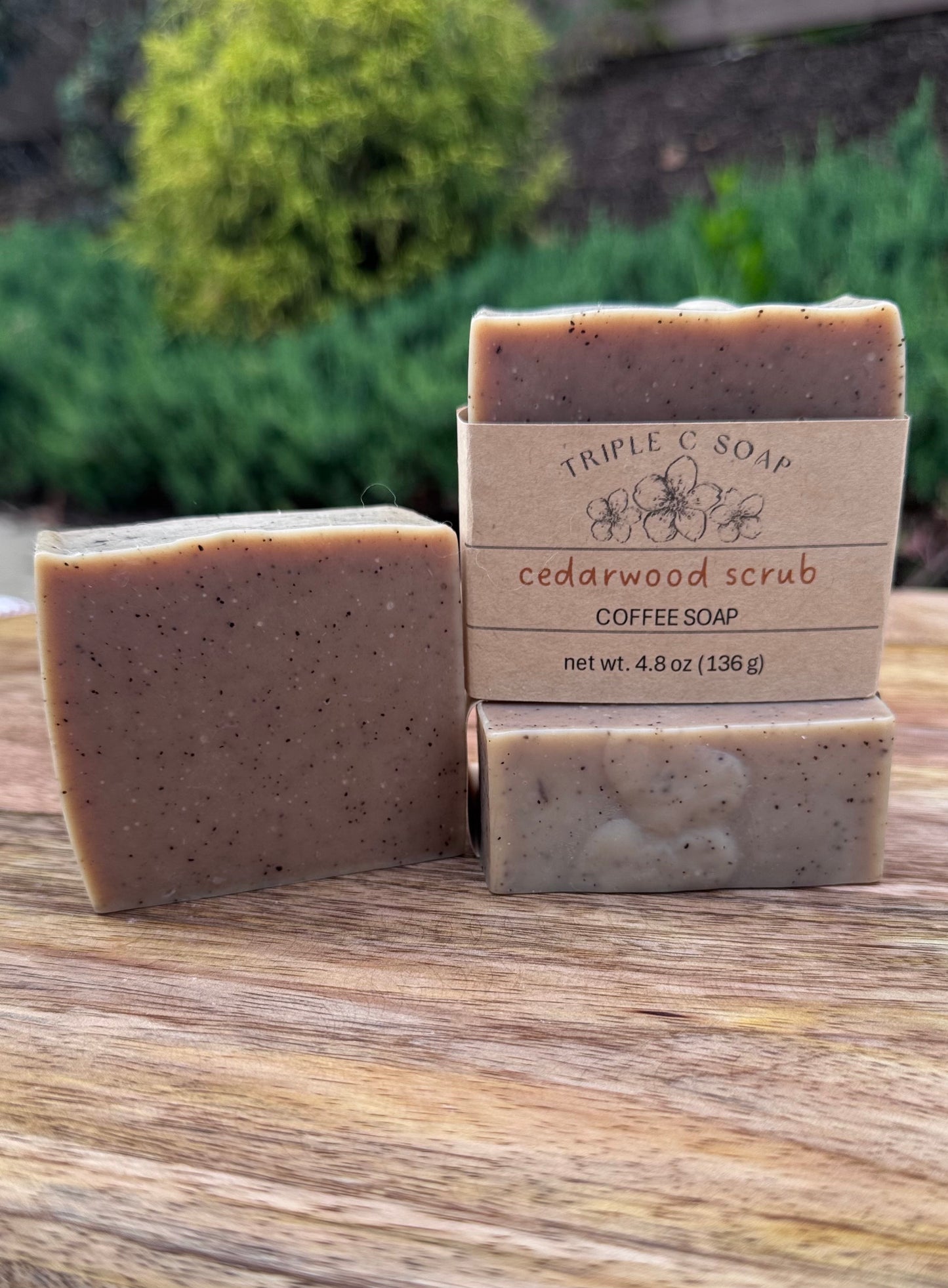 Three bars of cedarwood scrub soap on a wooden surface with a blurred green outdoor background.