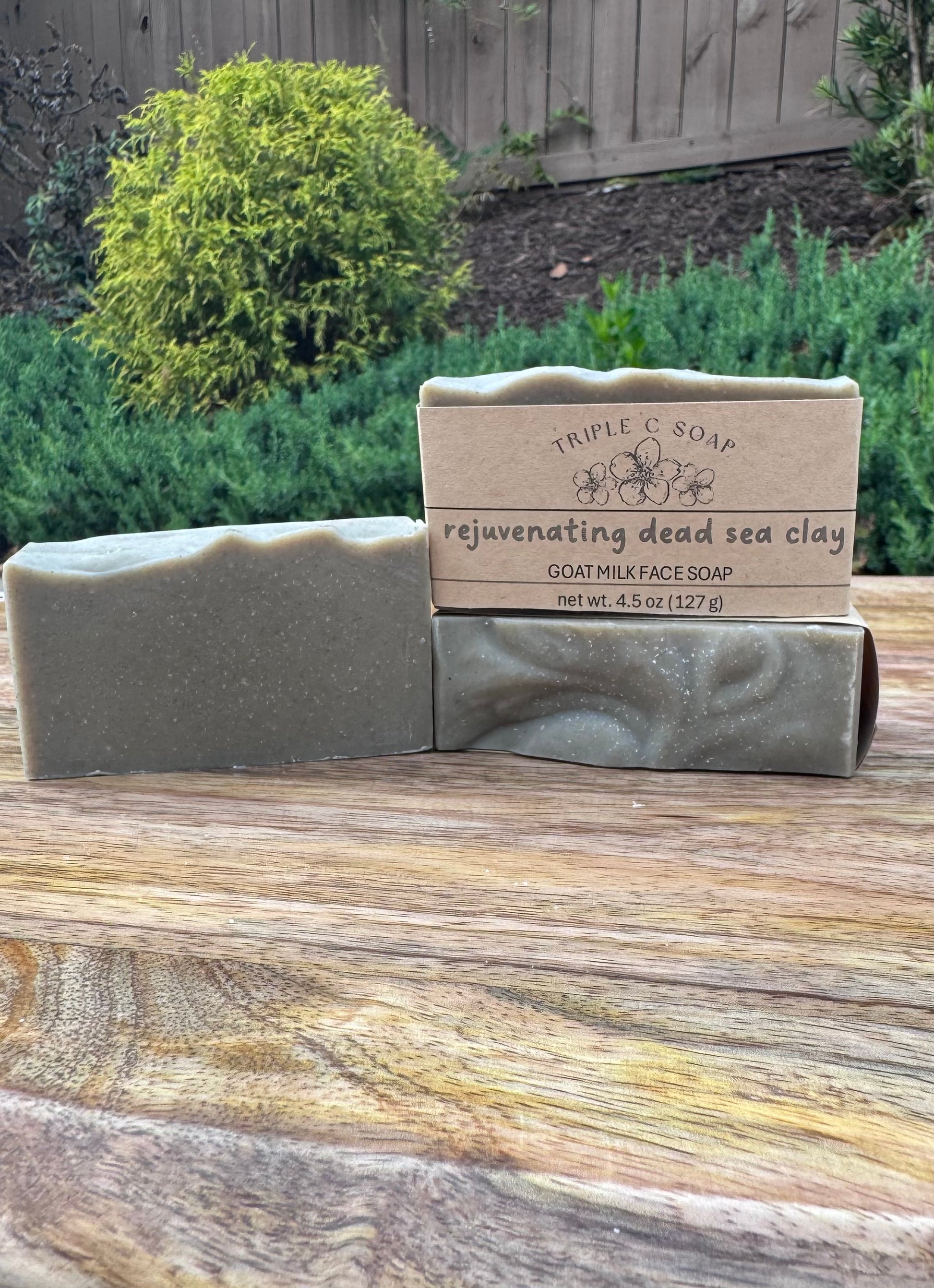 Three bars of gray soap with label 'rejuvenating dead sea clay' on wooden surface with natural background.