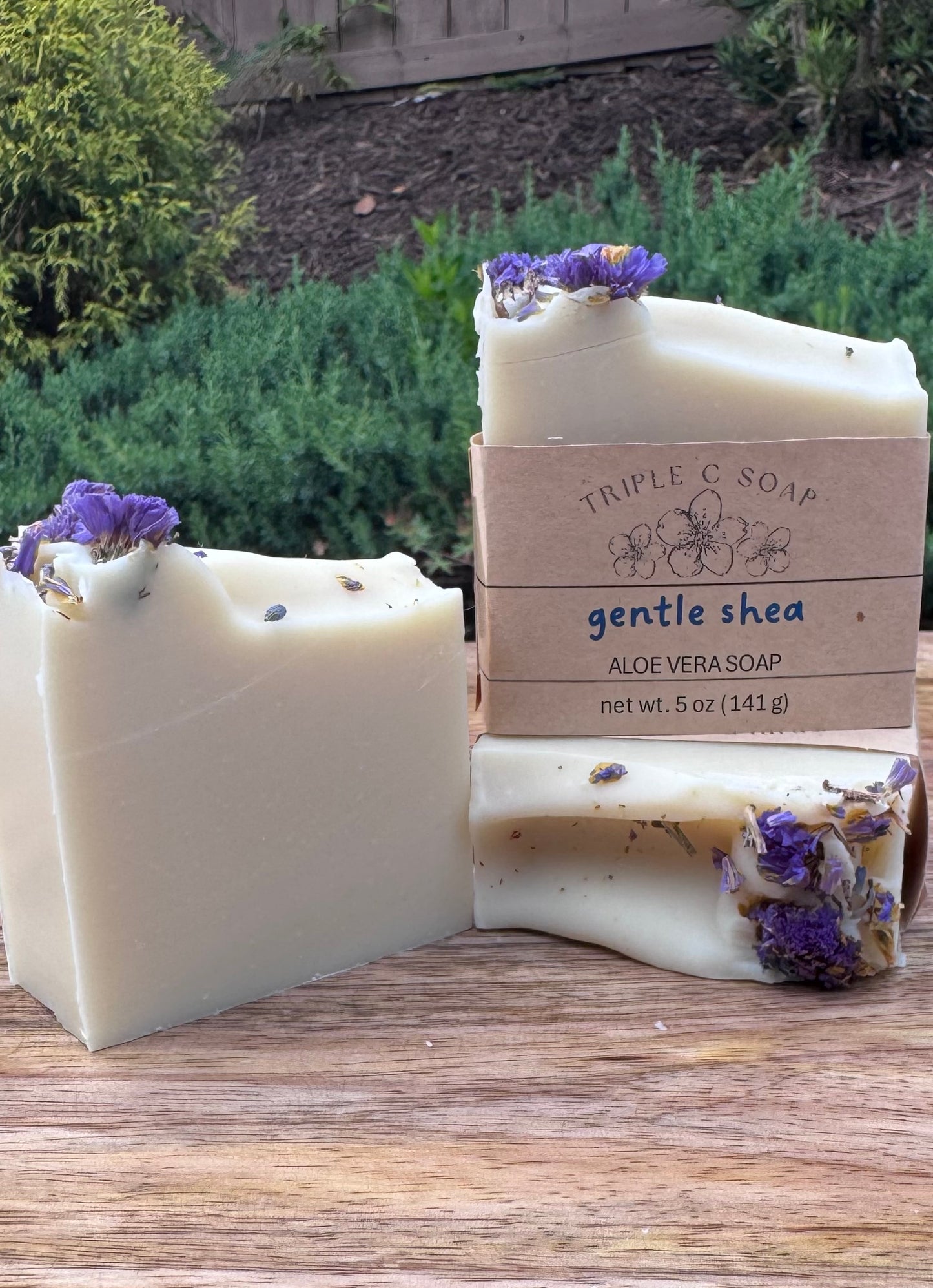 Three bars of aloe vera soap topped with forget-me-not flowers on a wooden surface, labeled 'Triple C Soap' in front of natural  background.