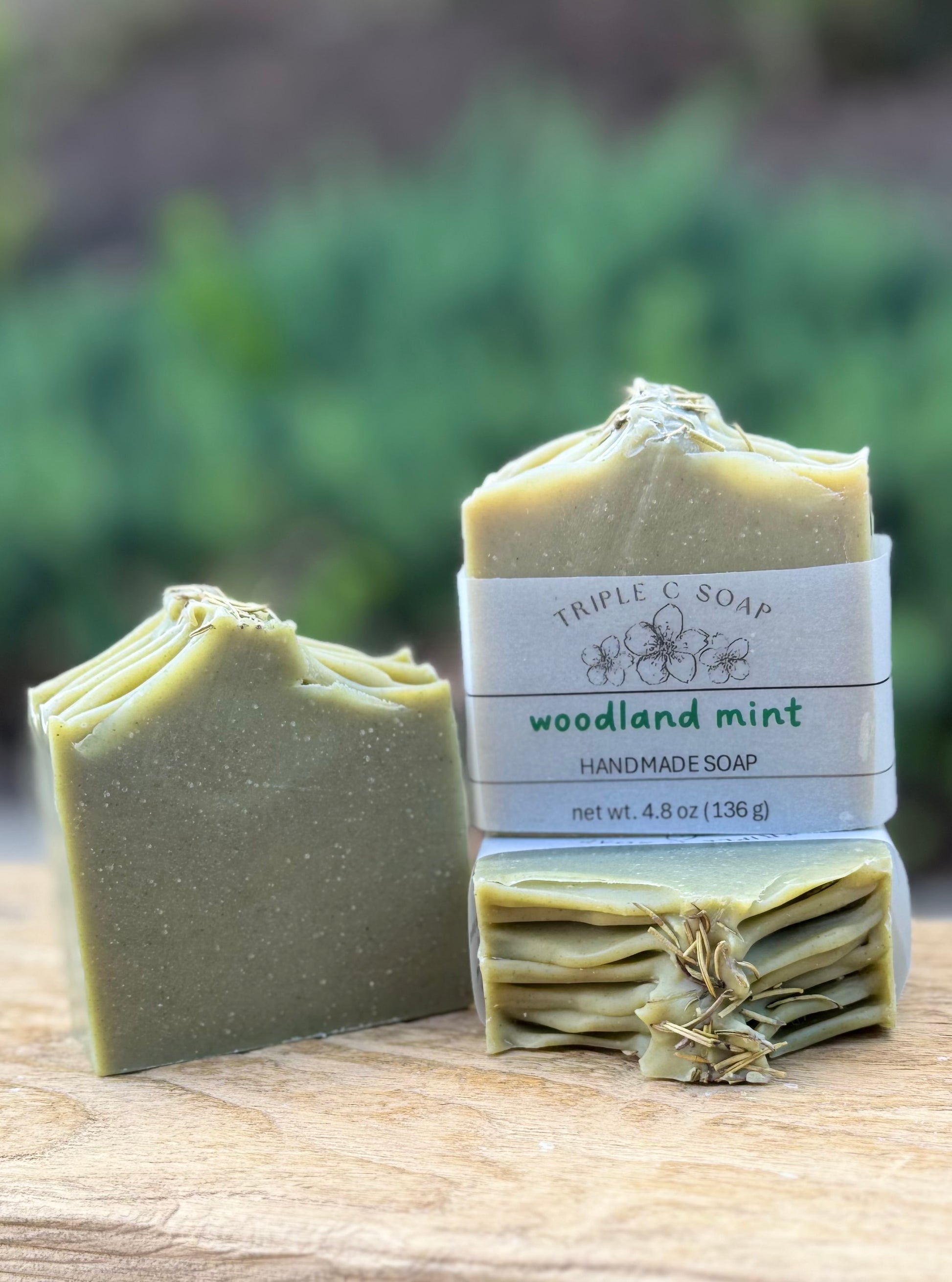 Handmade soap with packaging labeled 'Triple C Soap Woodland Mint' on a wooden surface with a blurred green background.
