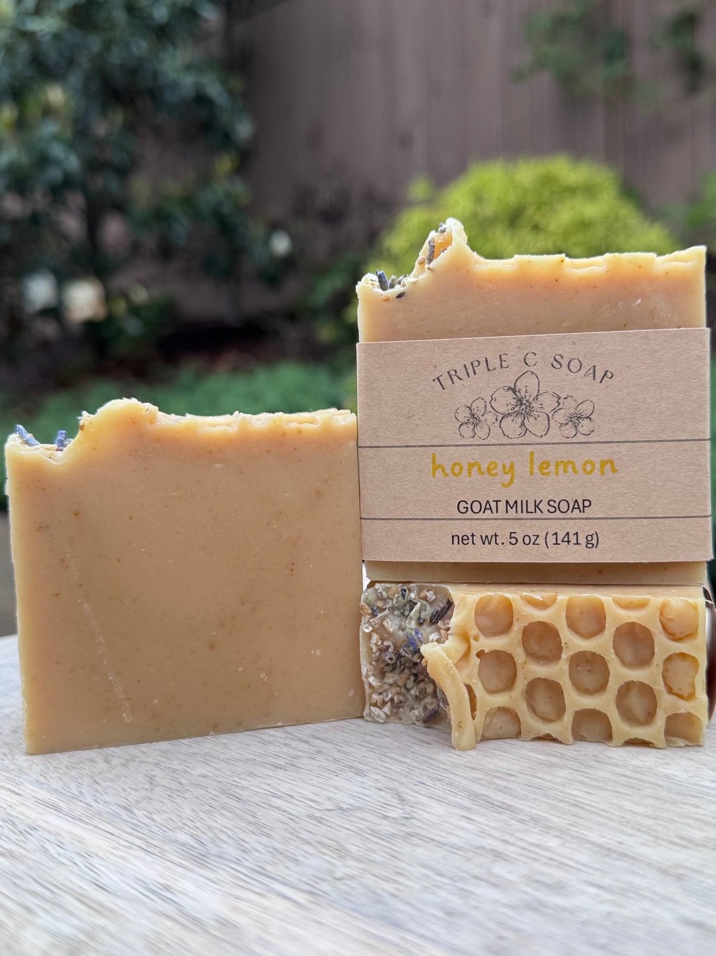 Three bars of Honey Lemon goat milk soap topped with dried lavender buds and fine cut oatmeal with a honeycomb pattern on a wooden surface outside.
