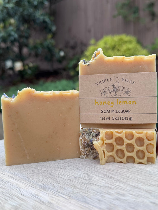 Three bars of Honey Lemon goat milk soap topped with dried lavender buds and fine cut oatmeal with a honeycomb pattern on a wooden surface outside.