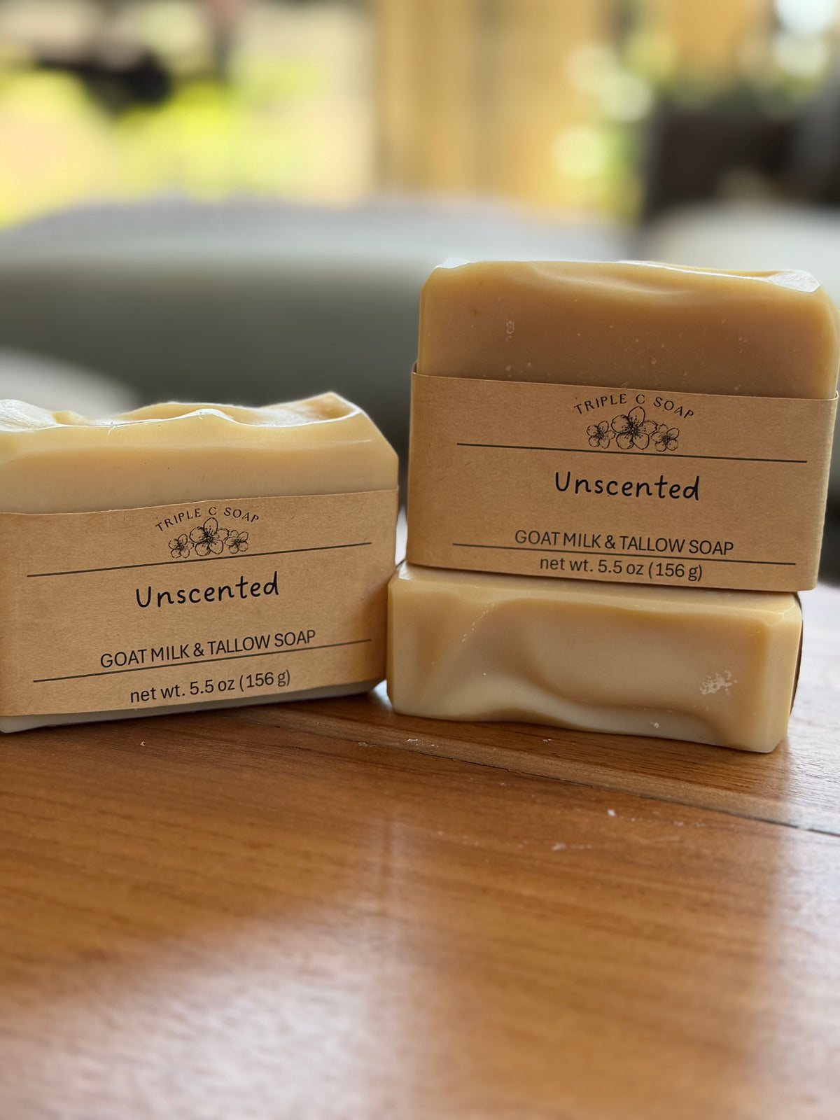 Three bars of Triple S Soap Unscented Goat Milk & Tallow Soap standing on a wooden table, with a softly blurred indoor background.